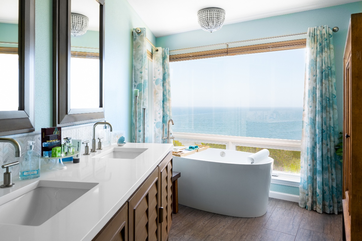 Stand alone tub in front of a floor to ceiling window with seafoam blue/green curtains drawn open