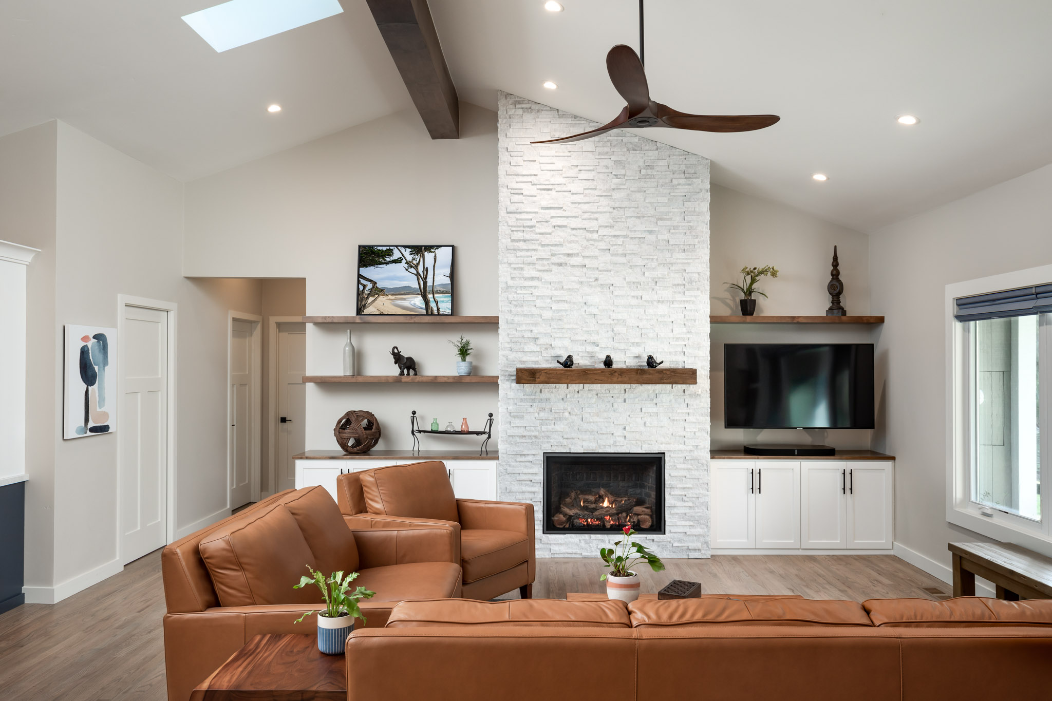 Brown leather sectional sitting in a spacious living room with a vaulted ceiling, integrated fireplace, and wooden floating shelves