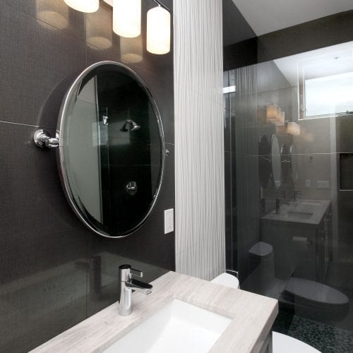 Bathroom sink with black tile walls, grey marbled countertop, and a round mirror